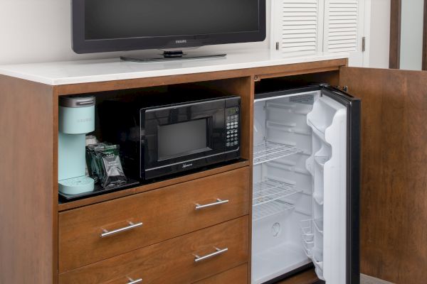 The image shows a wooden cabinet with an open mini-fridge, a microwave, and a coffee maker inside. A TV is on top of the cabinet.