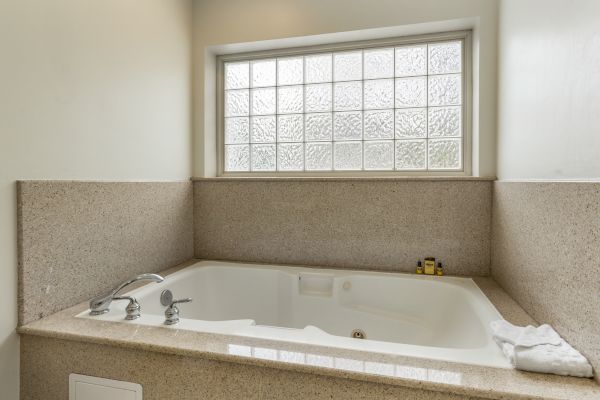 This image shows a modern bathroom with a large bathtub, frosted glass window, and light-colored countertops and walls. A towel and toiletries are visible.