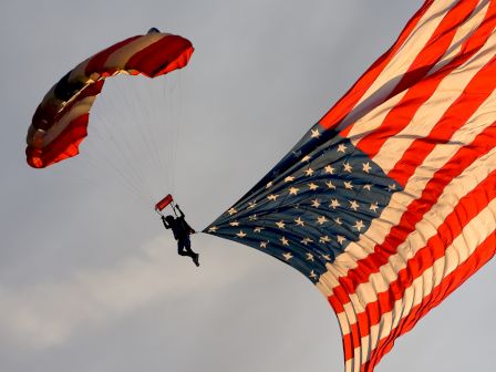 A skydiver is descending with a large American flag, showcasing a patriotic display in the sky, with their parachute open against a clear backdrop.