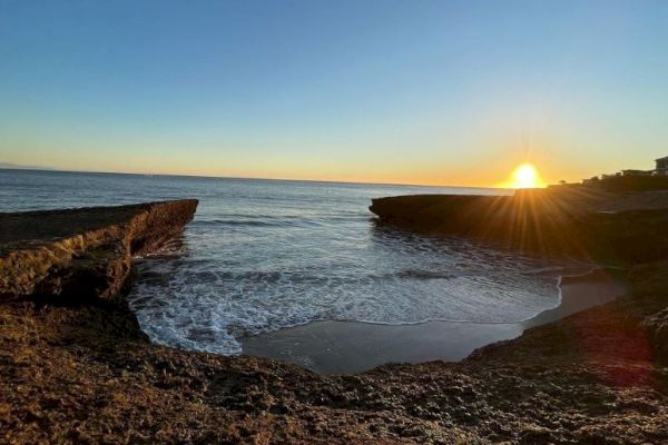 The image shows a beautiful coastal scene at sunset with rocky cliffs framing the water and the sun setting on the horizon, casting a golden glow.
