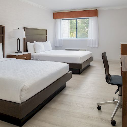 The image shows a hotel room with two double beds, a desk, and a chair, with a window providing natural light.