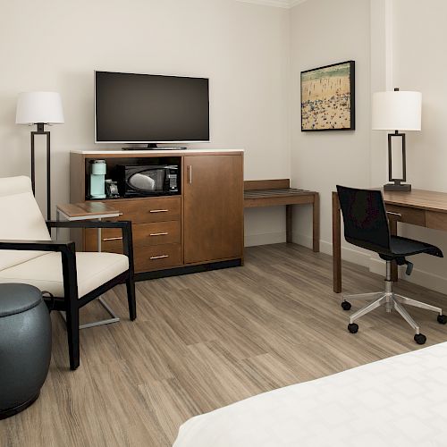 This image shows a modern hotel room with a TV, dresser, desk, lamps, chair, and white bedding. The floors are wooden.