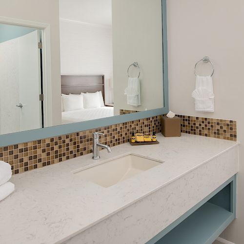 A modern bathroom with a large mirror, sink, and a neatly folded towel, adjacent to a bedroom and a small office desk.
