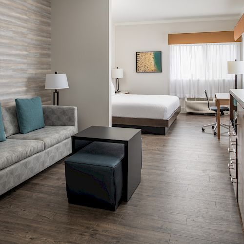 A modern hotel room with a sofa, ottomans, TV stand, lamp, bed, work desk, and floor lamp, featuring neutral and blue accents, and wooden flooring.