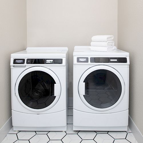 The image shows a laundry room with a washer and dryer side by side, both white. There are neatly folded white towels on top of the dryer.