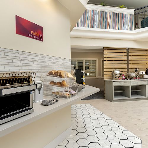 The image shows a breakfast area with a toaster oven, bread and pastries on trays, adjacent counters with beverages, and a modern design.