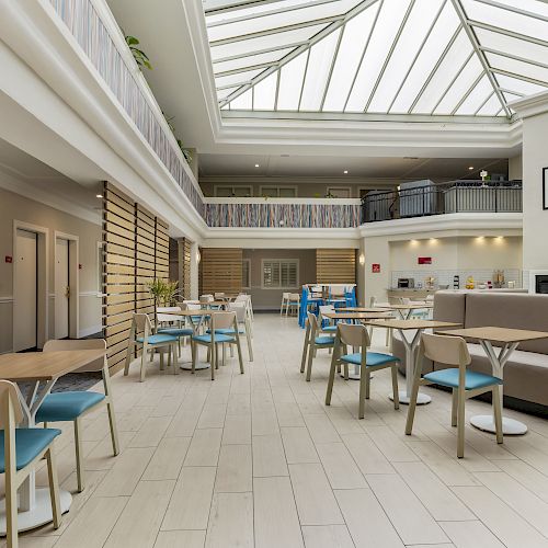 A modern, well-lit atrium with numerous tables and chairs, skylight ceiling, and a cozy seating area surrounded by wooden accents and modern decor.