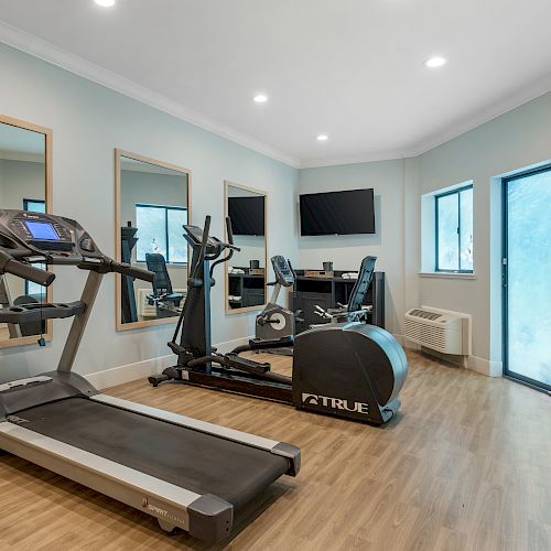 A home gym with a treadmill, exercise bike, and elliptical trainer. There are mirrors, a TV, and windows on the light-colored walls ending the sentence.