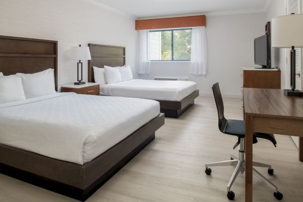 The image shows a clean, modern hotel room with two double beds, a desk with a chair, a TV, and a window with curtains.