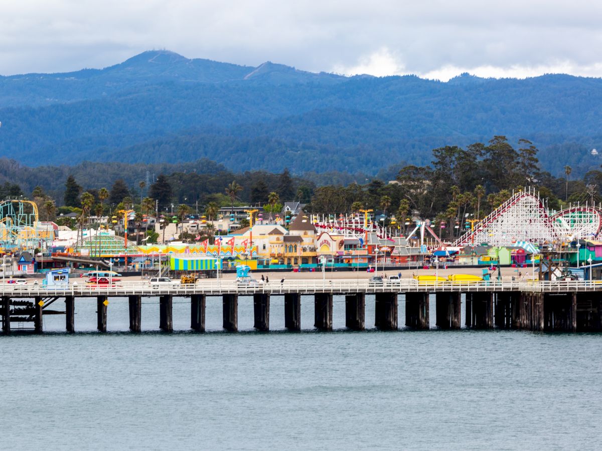 An amusement park beside a large body of water with various rides, bright colors, and mountains in the background under a partly cloudy sky.